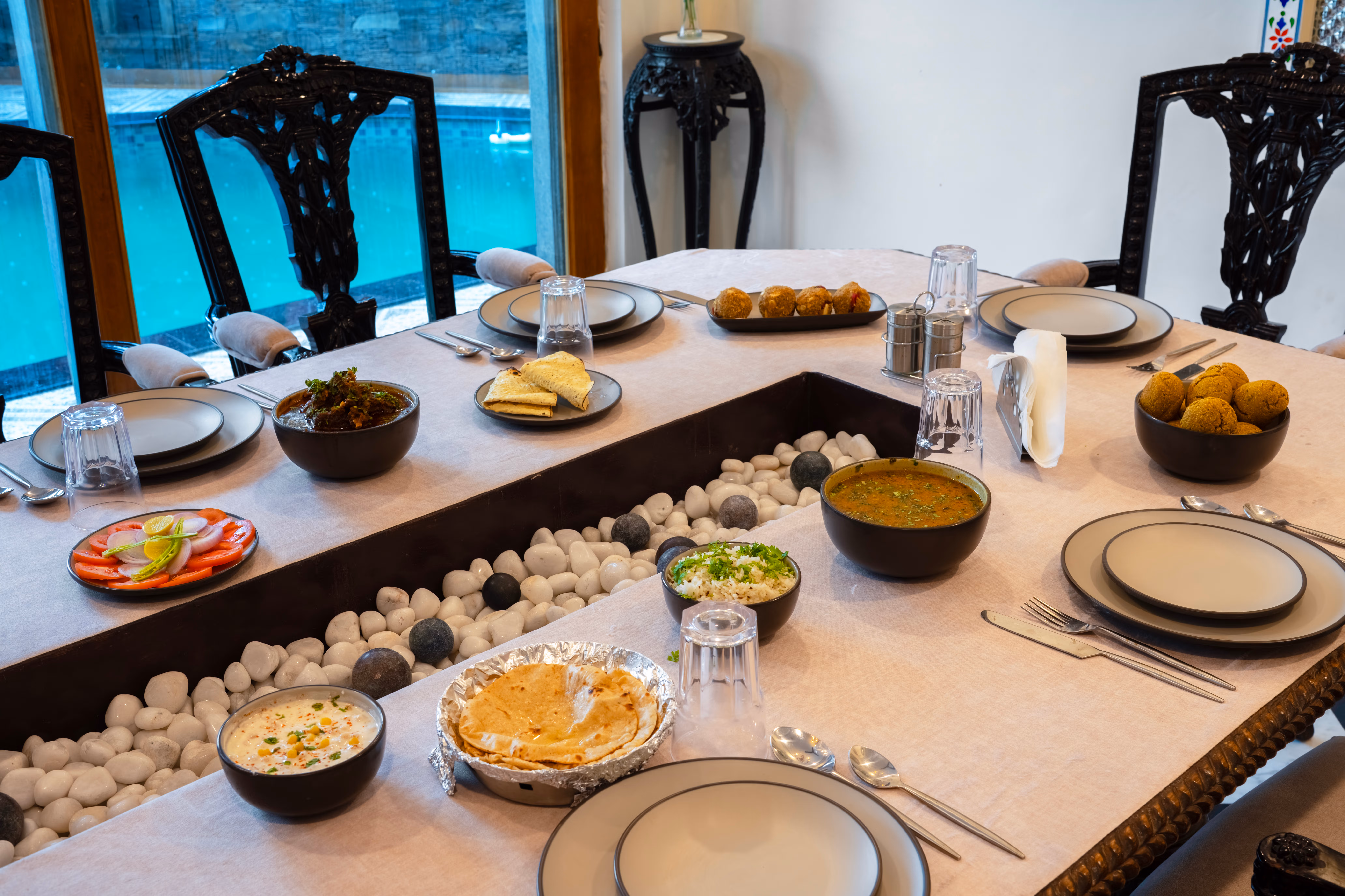 Dining table laid with plates, glasses and serving dishes beside carved wooden chairs and large windows.