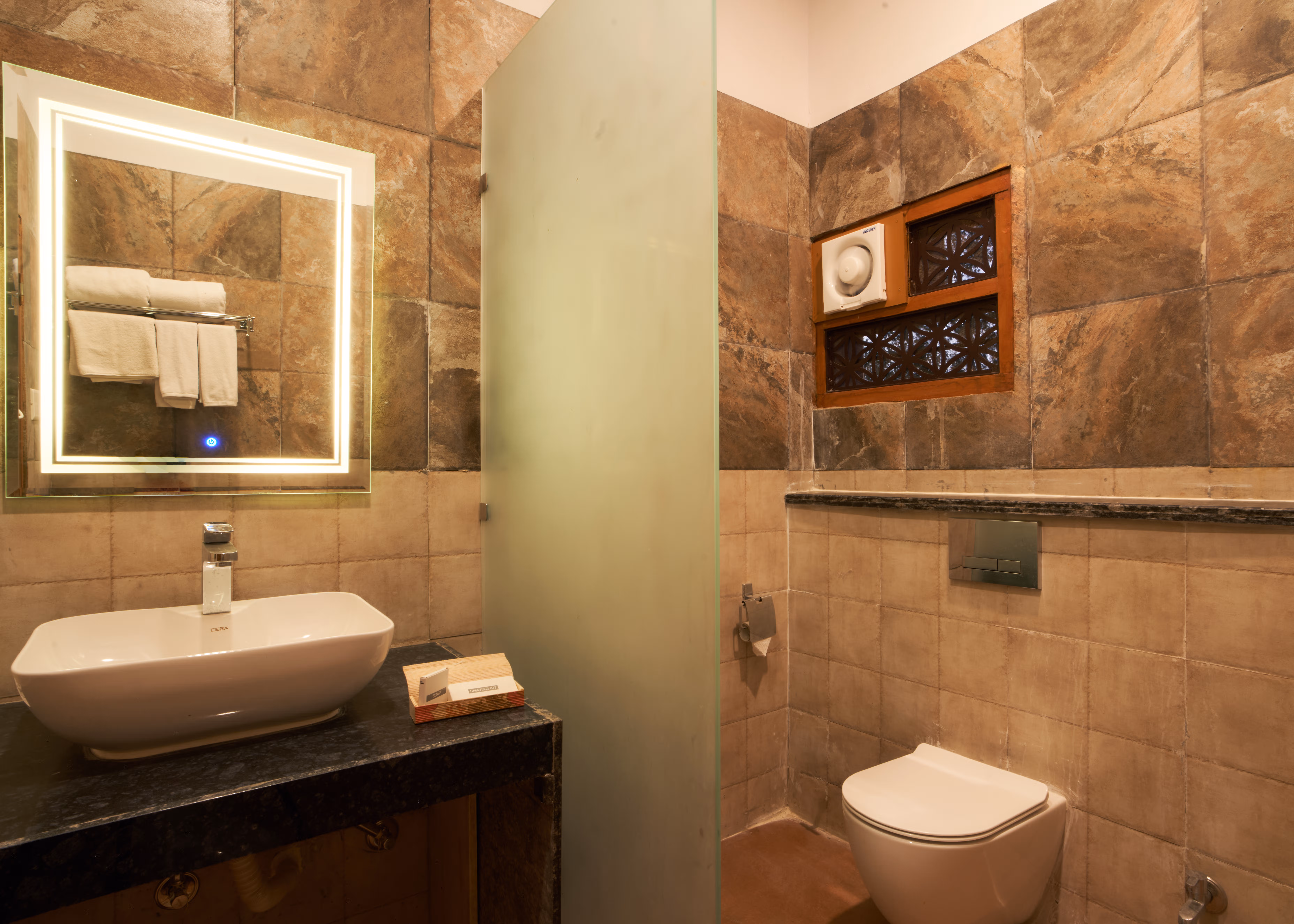 Stone-tiled bathroom with a countertop basin, illuminated mirror, frosted shower partition and wall-mounted toilet.