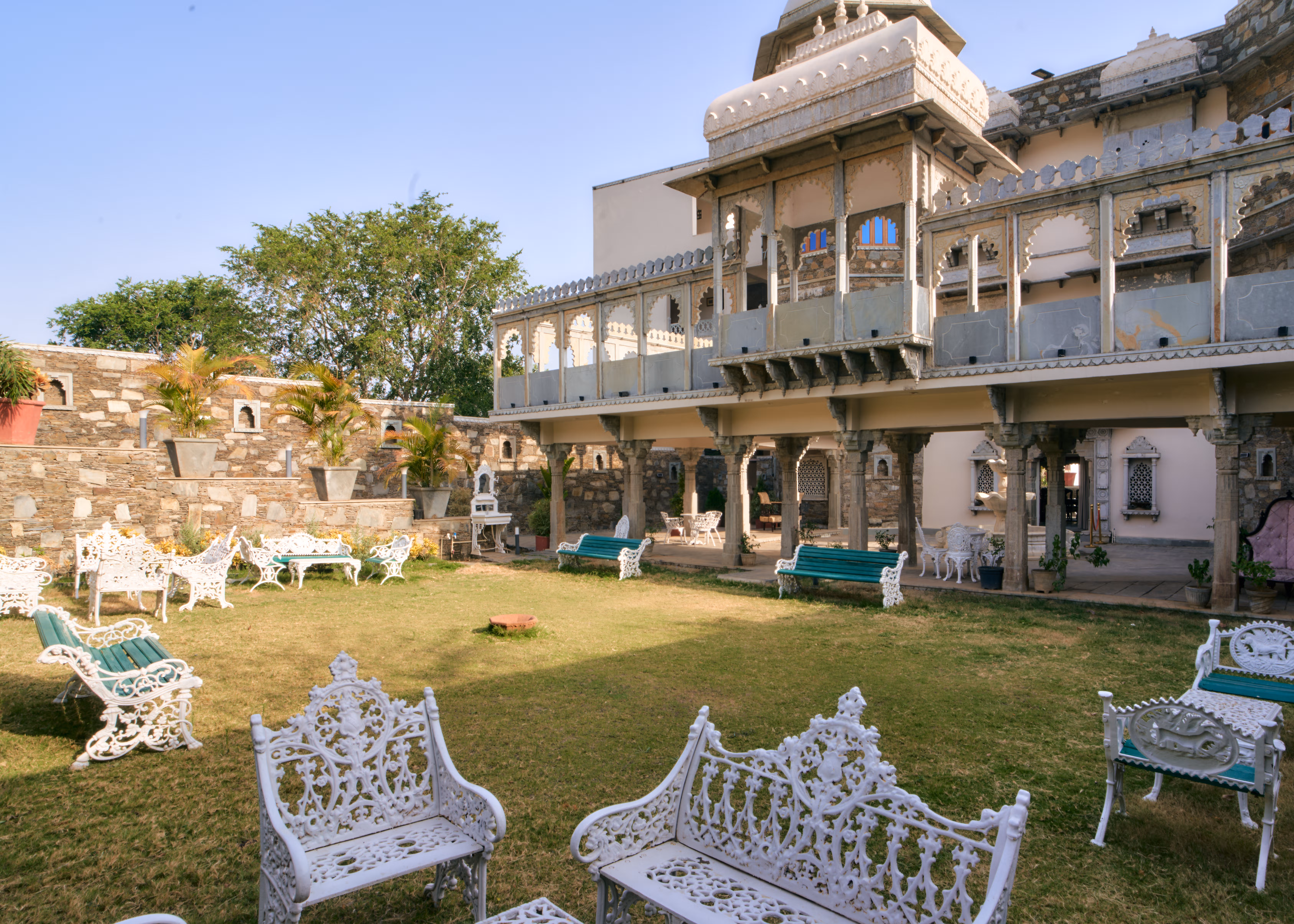 Palace lawn with white metal garden furniture, green benches and a two-storey building with carved verandahs.