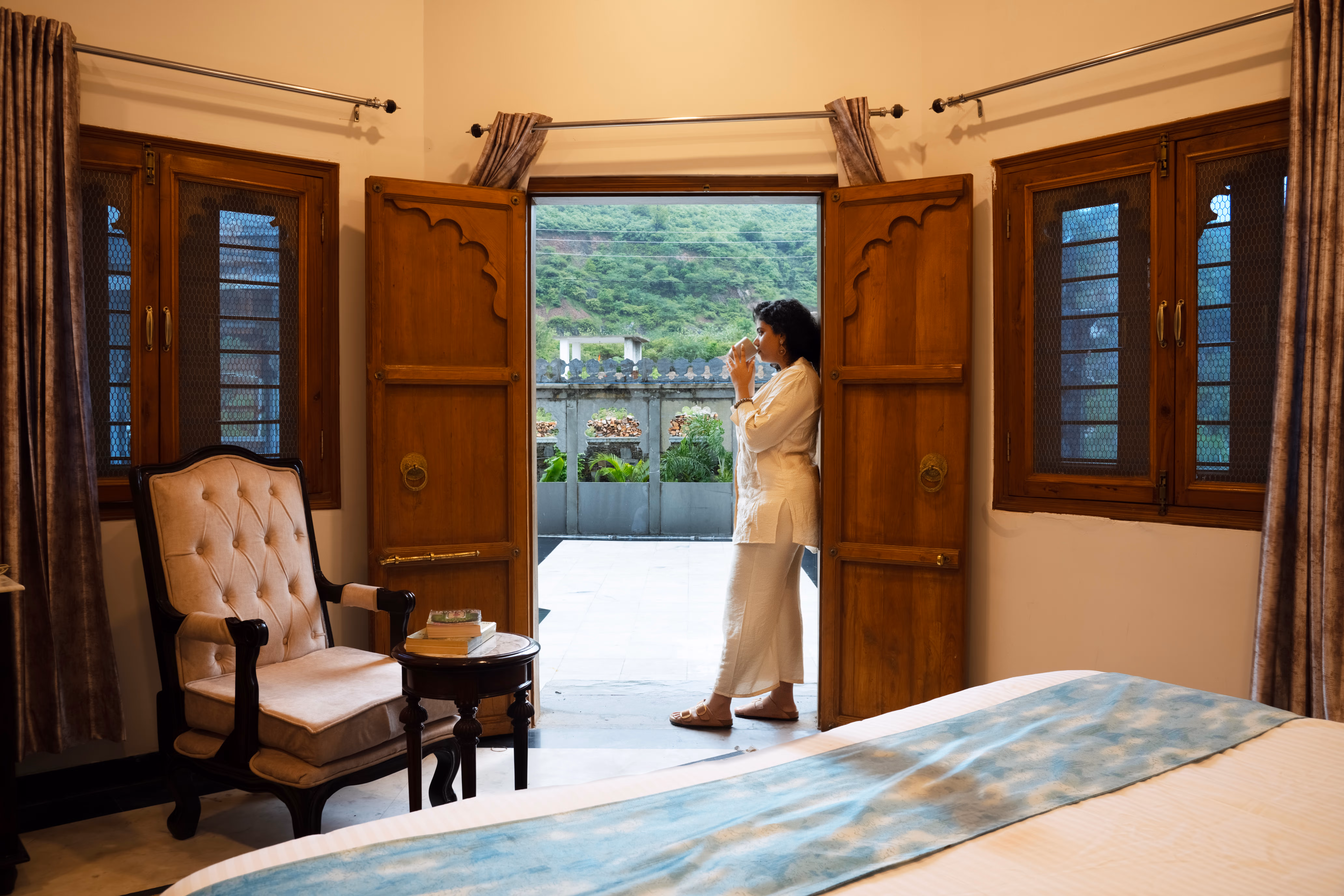 Guest standing beside open wooden bedroom doors leading to a balcony overlooking green hills.
