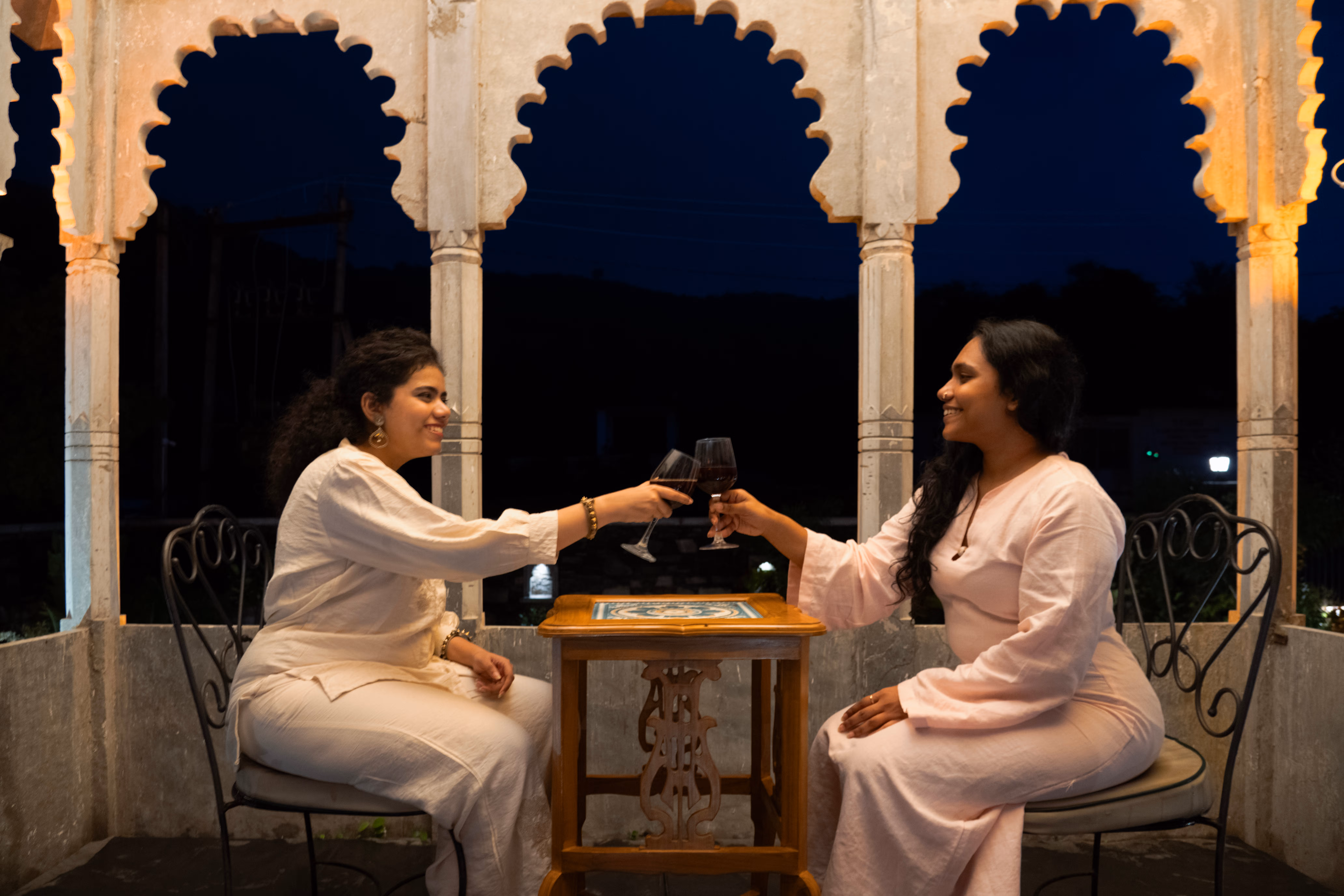 Two guests raising glasses at a small table beneath illuminated stone arches at night.