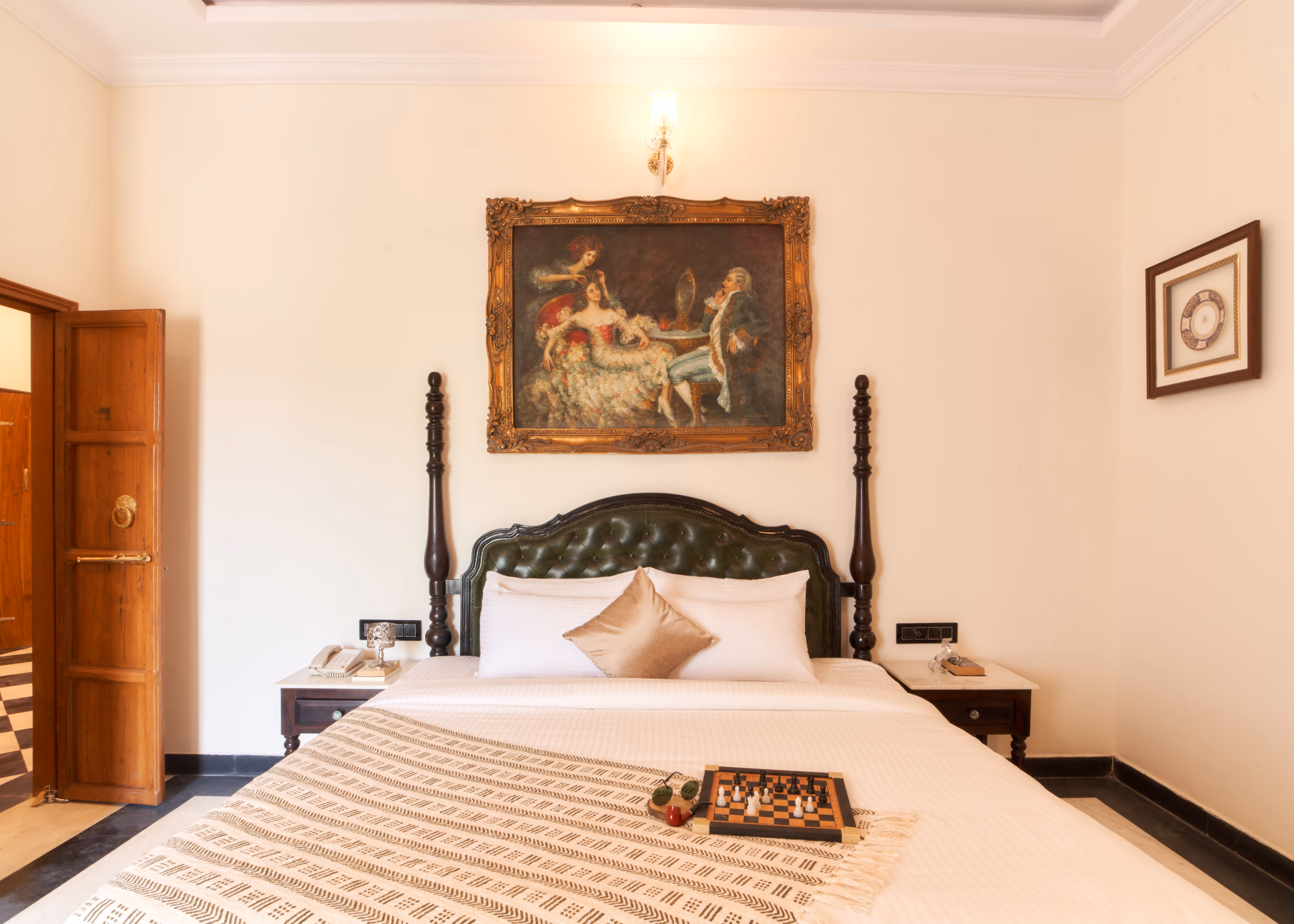 Bed with a carved wooden headboard, white linen and a striped runner beneath a framed painting.