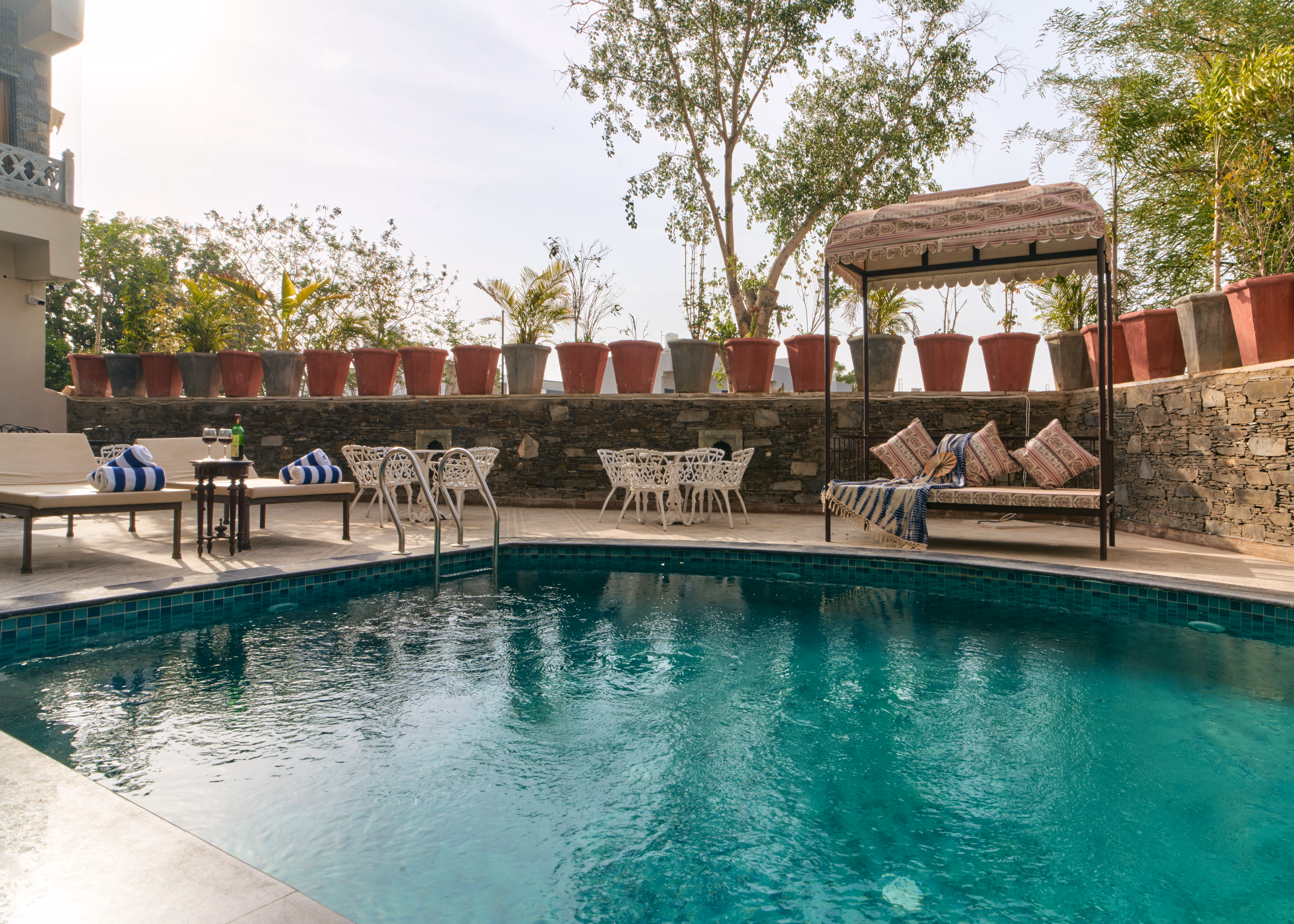 Swimming pool bordered by stone paving, white garden furniture, potted plants and shaded cushioned seating.