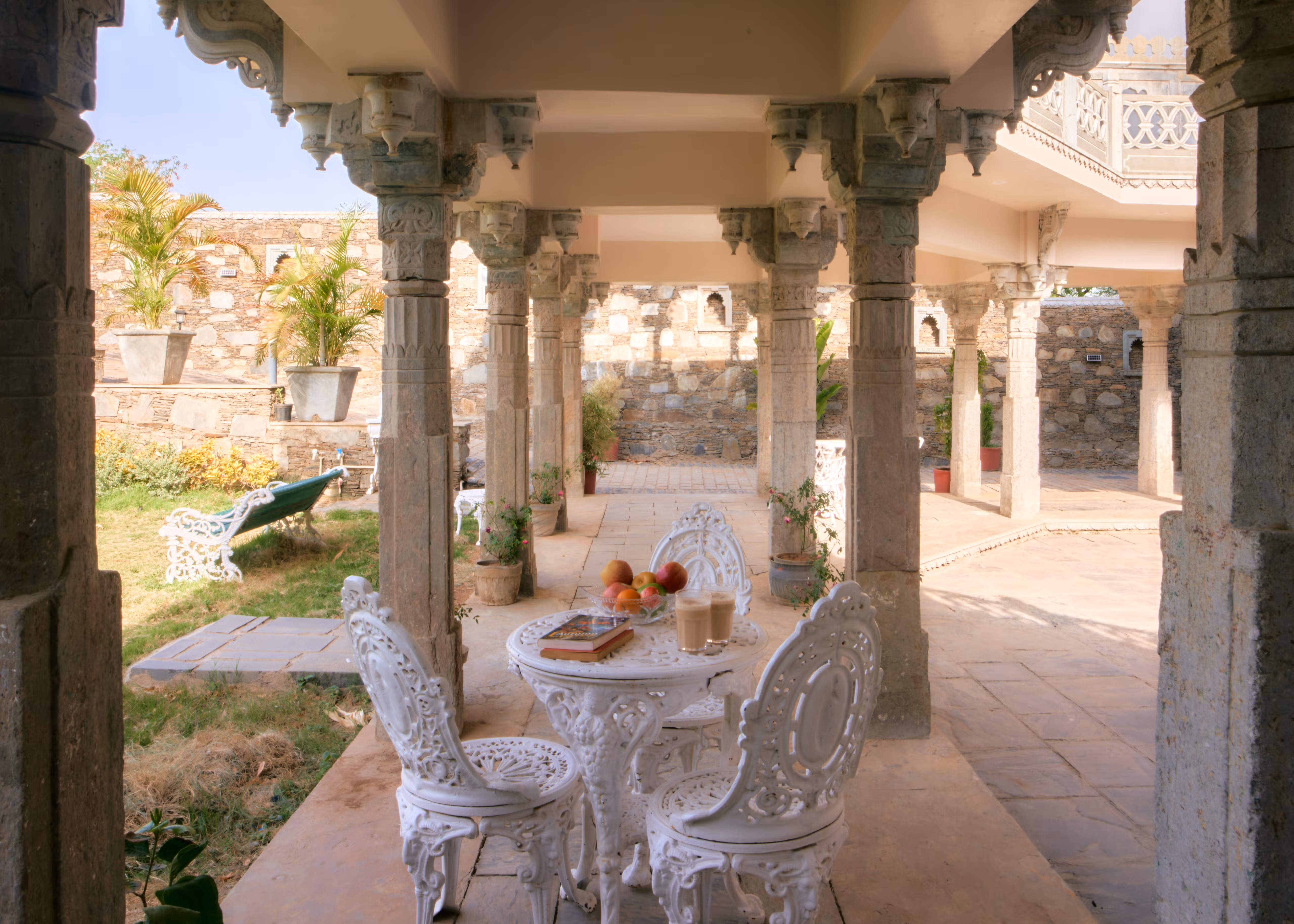 White outdoor table and chairs with a refreshment tray beneath a stone colonnade beside the lawn.