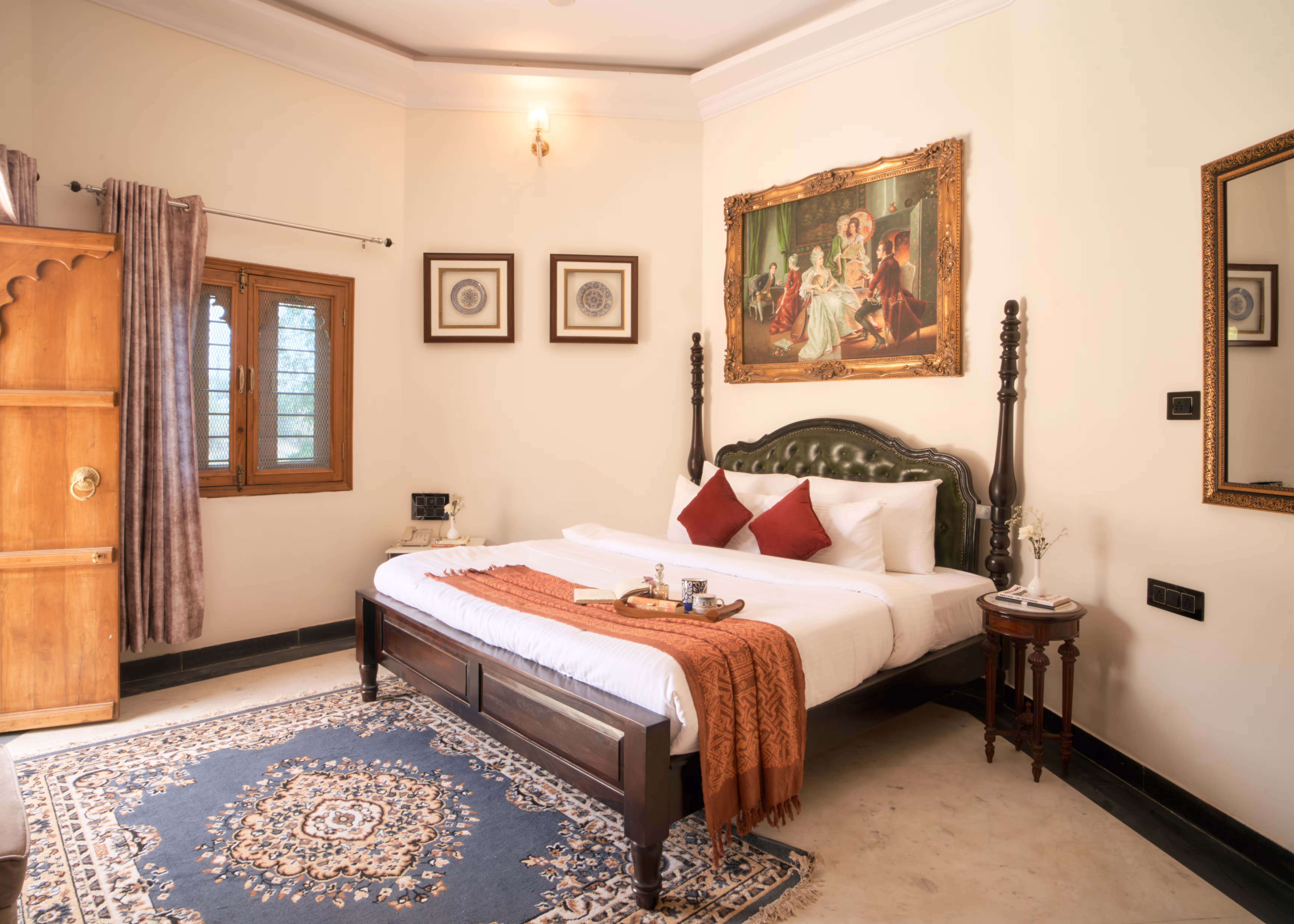 Bedroom with red cushions, a rust-coloured runner, carved wooden furniture and open doors admitting daylight.
