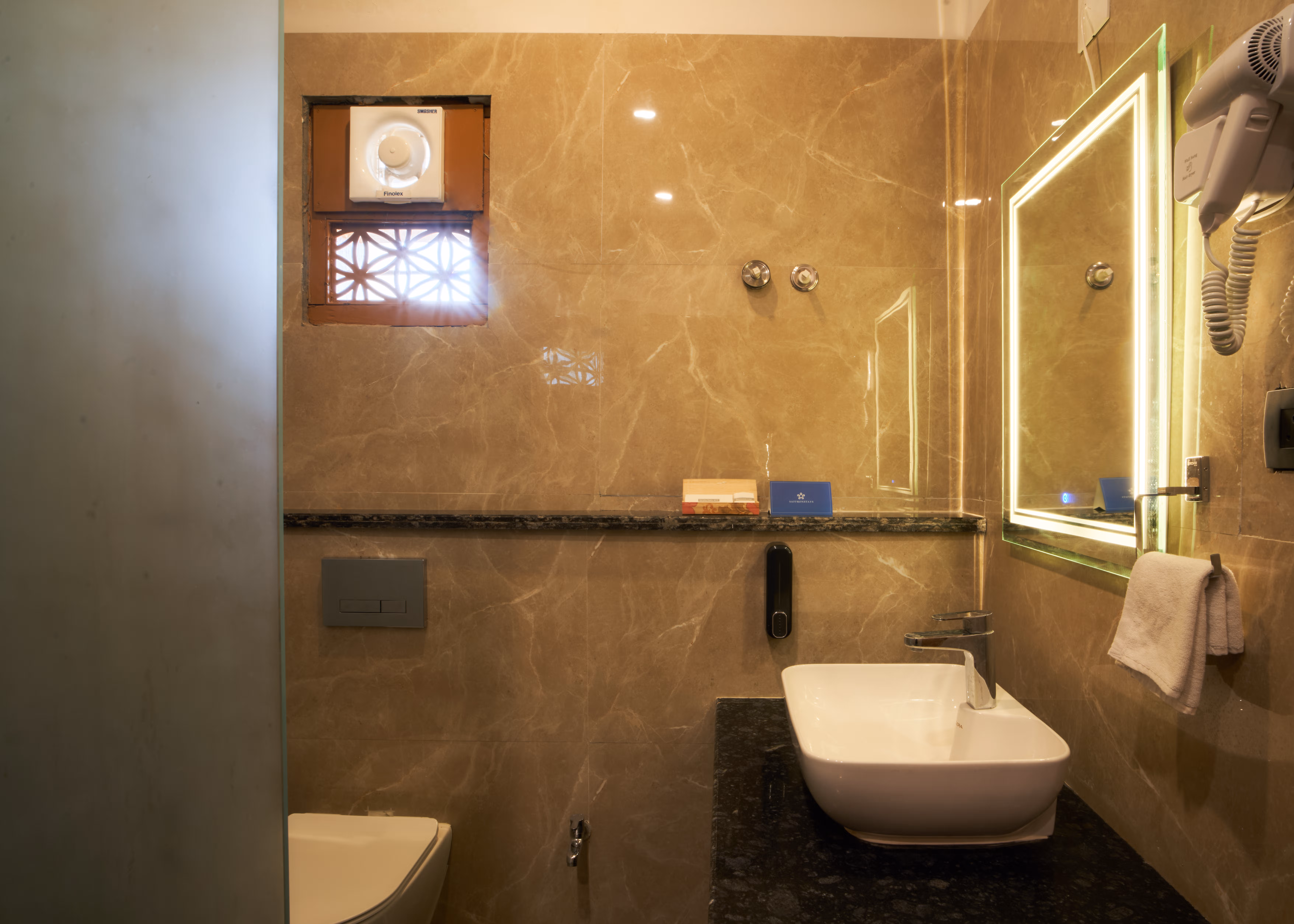Bathroom with warm-toned stone tiles, a white basin, illuminated mirror, towel rail and glass shower enclosure.