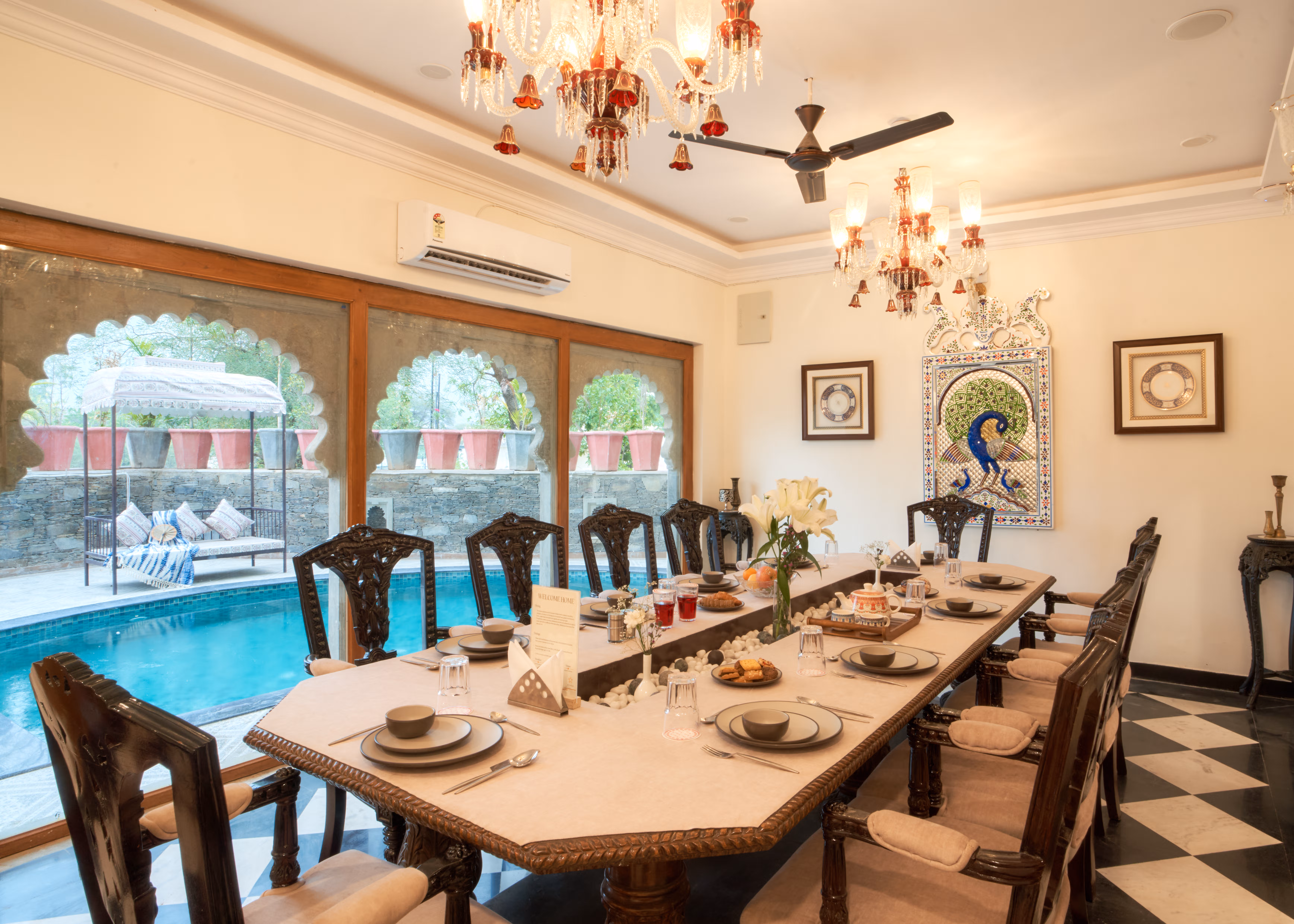 Long dining table set with crockery beneath a red-accented chandelier, beside arched garden-facing windows.