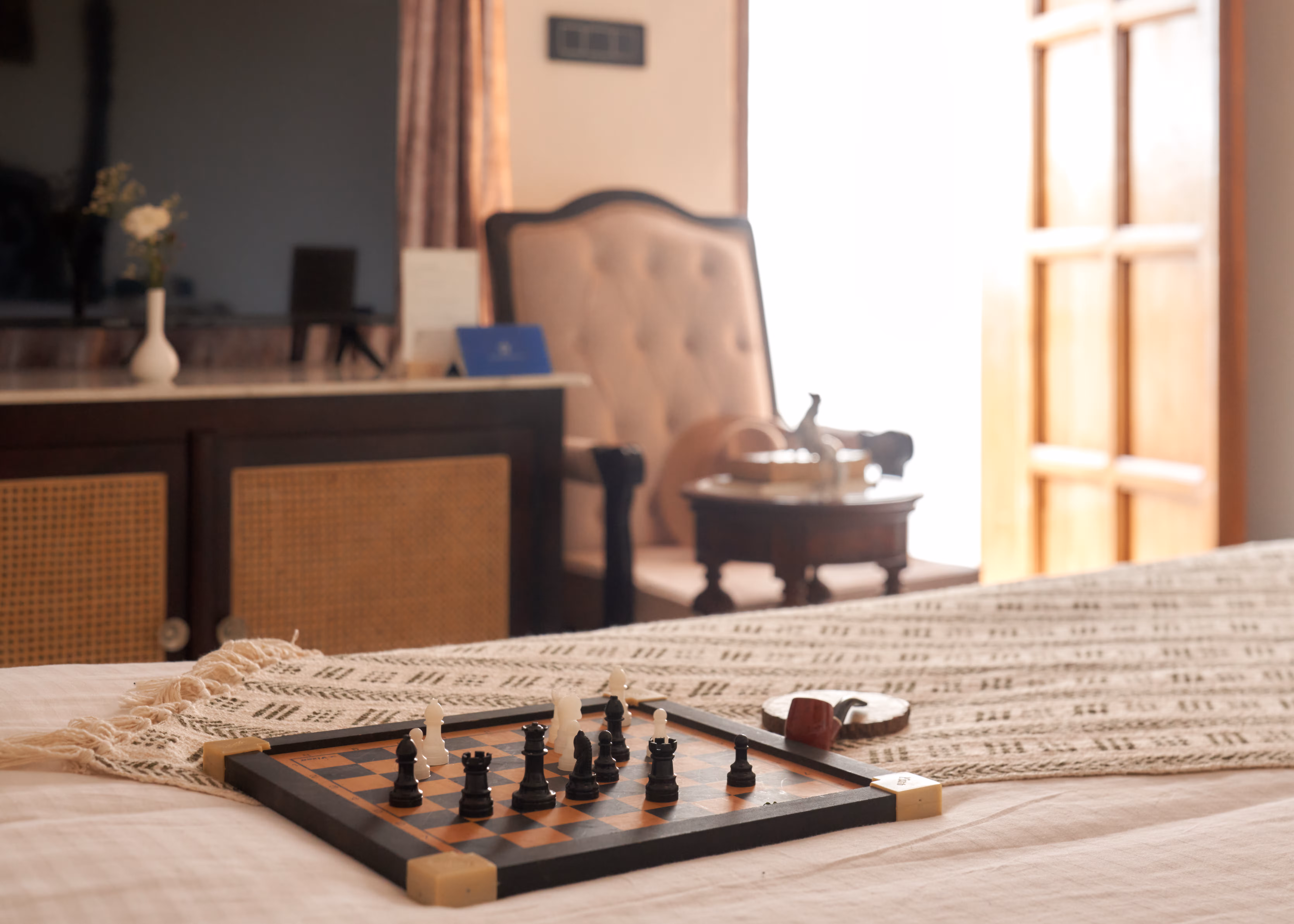 Chessboard placed on a patterned bed runner, with an upholstered chair and wooden furniture in the background.