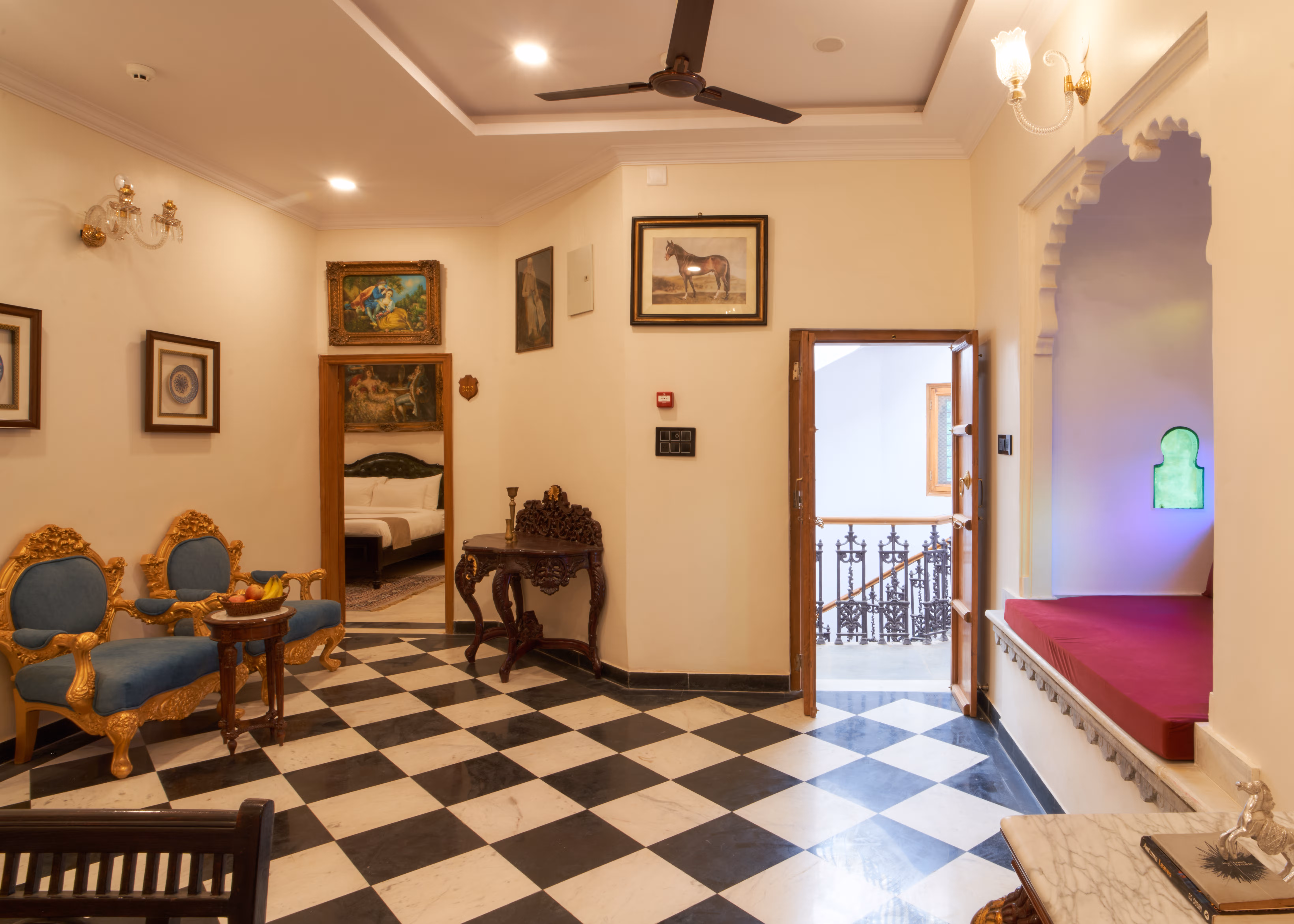 Sitting area with blue upholstered chairs, framed pictures, a magenta window seat and an open balcony doorway.