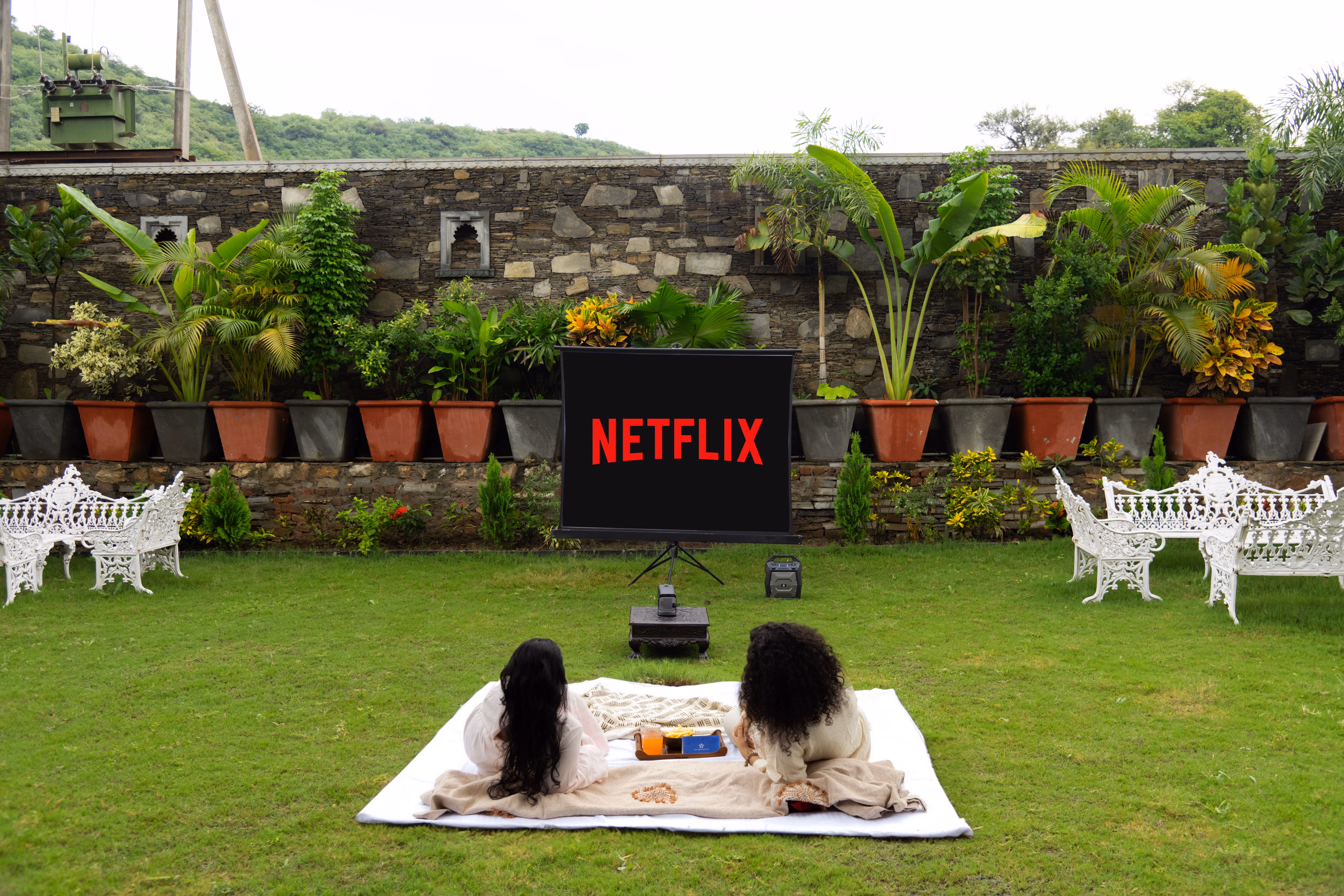 Guests reclining on the lawn facing an outdoor movie screen, surrounded by garden seating and potted plants.