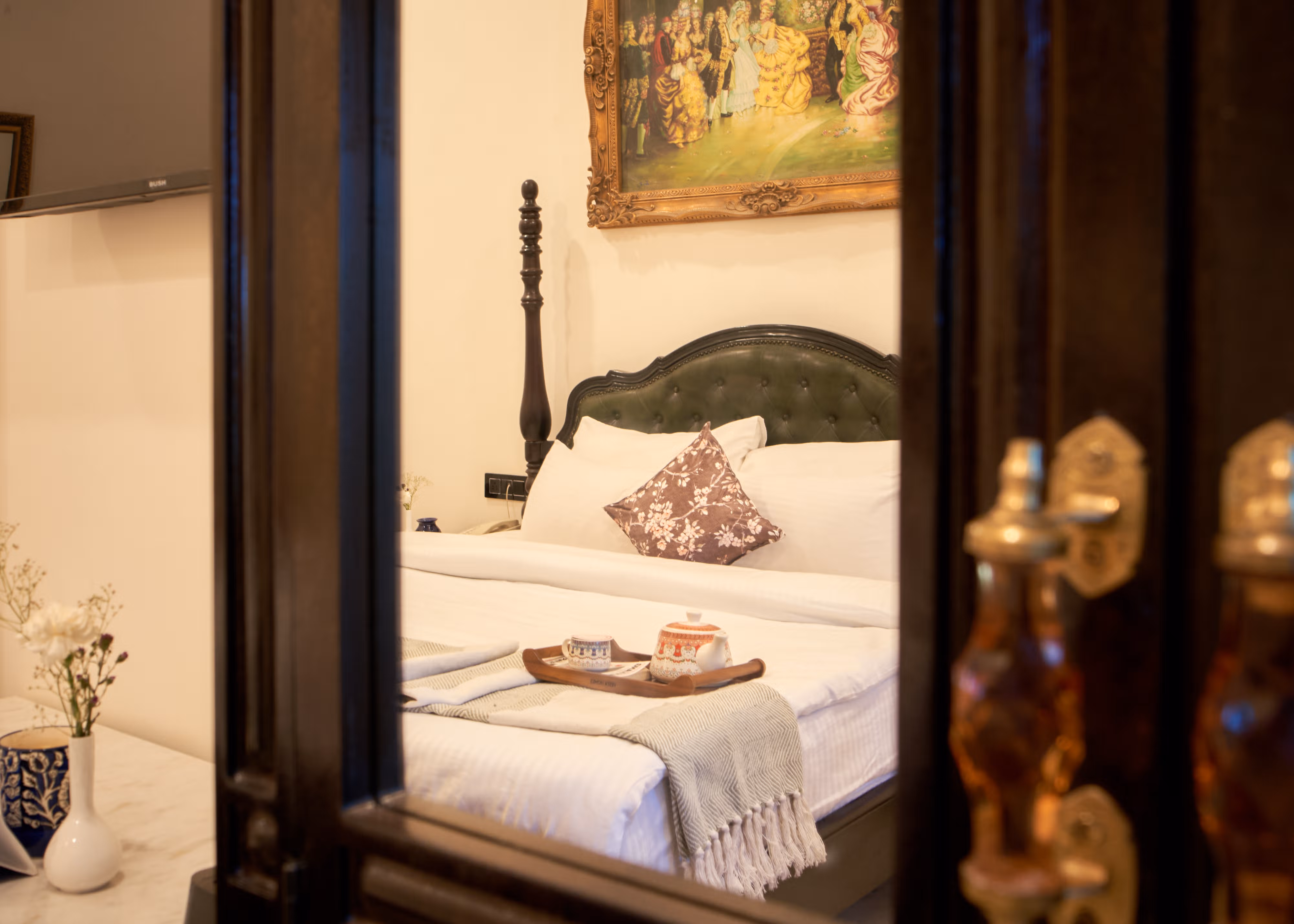 View through a dark wooden doorway towards a bed with white linen, a patterned cushion and a refreshment tray.
