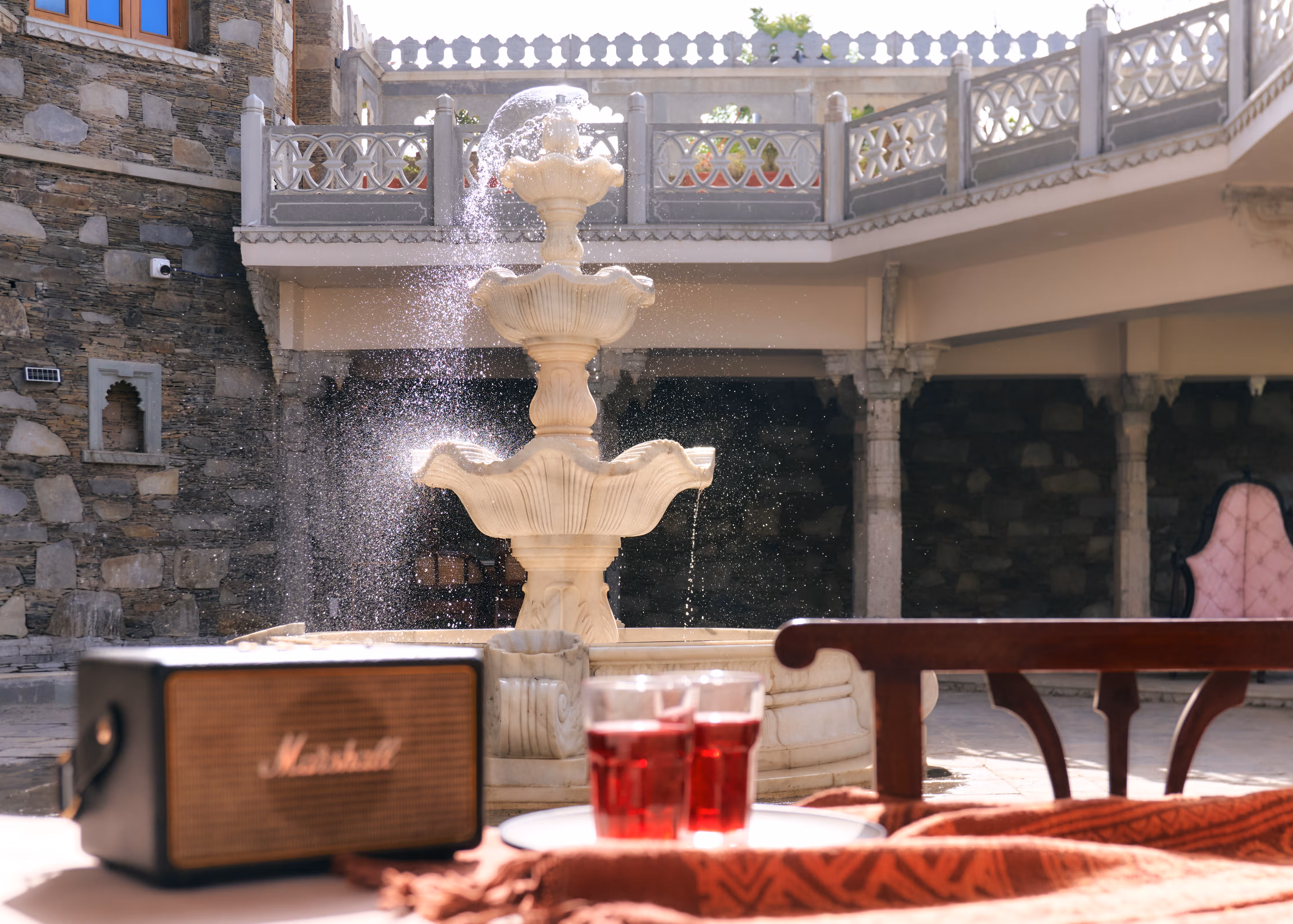 Table with a speaker and red drinks beside a flowing courtyard fountain and carved stone balcony.
