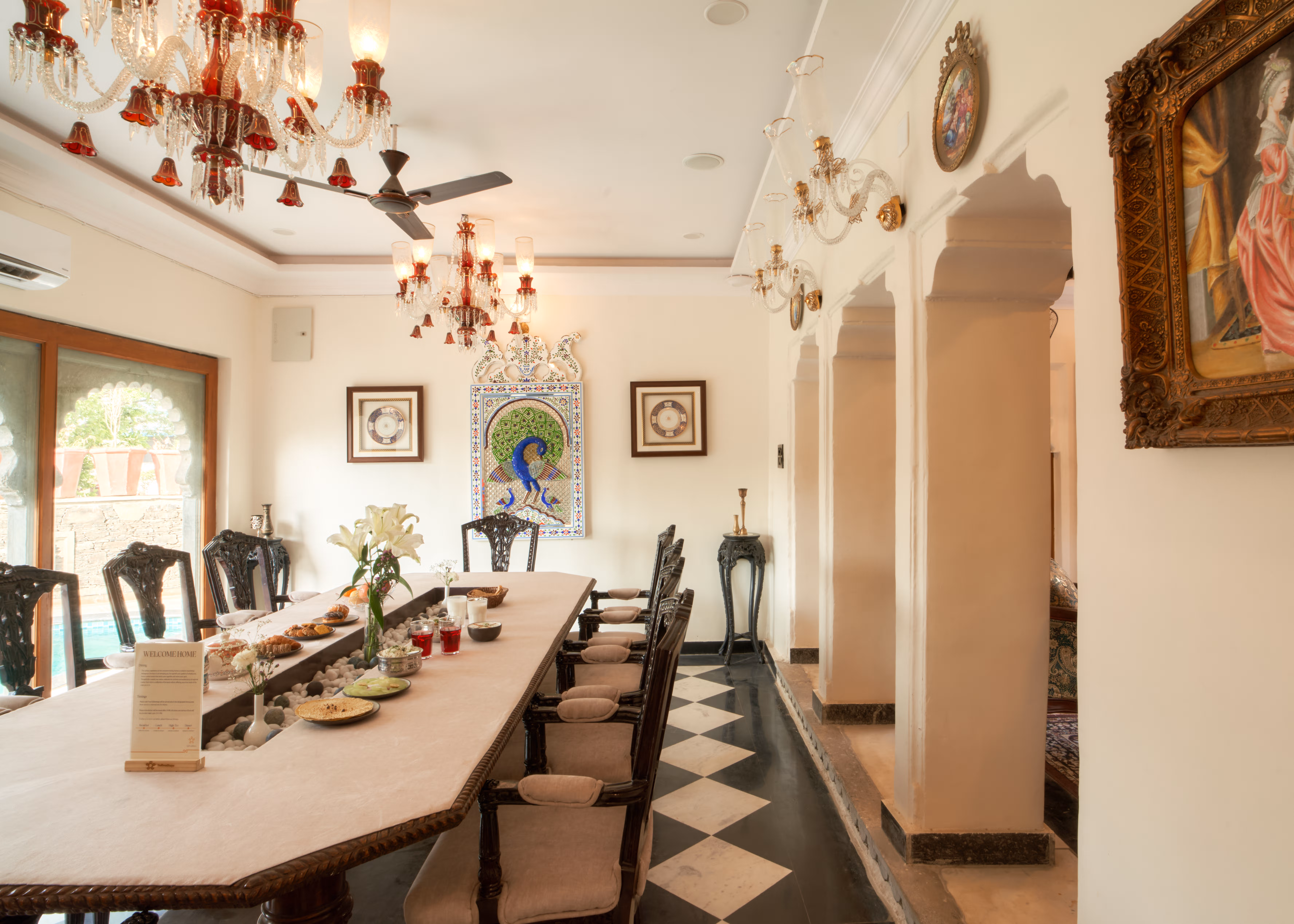 Long dining table with carved chairs, crockery and a floral centrepiece beneath a chandelier.