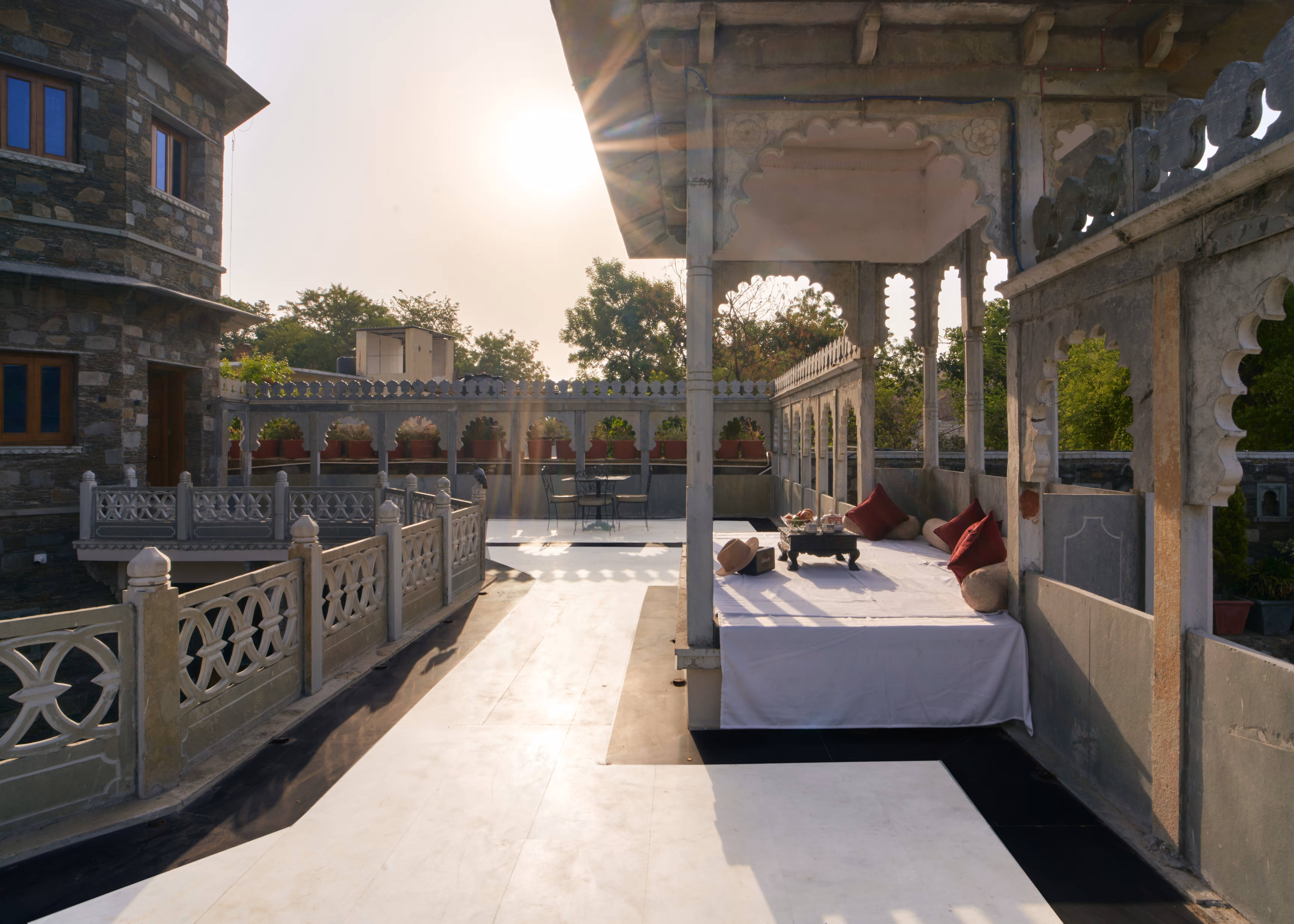 Cushioned daybed beneath a carved stone pavilion beside a sunlit terrace and decorative balustrades.