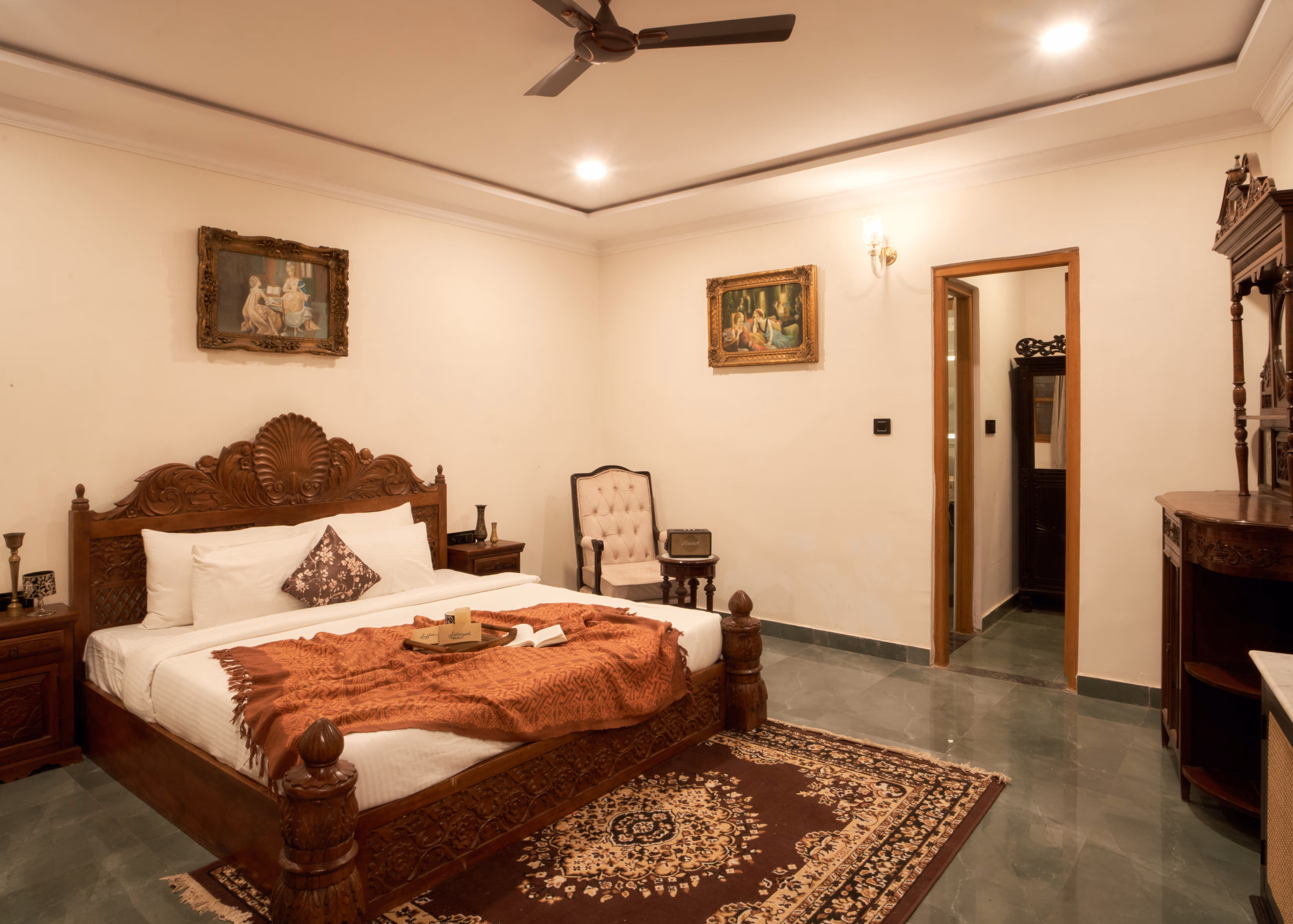 Bedroom with a carved wooden bed, rust-coloured runner, patterned rug, upholstered chair and framed paintings.