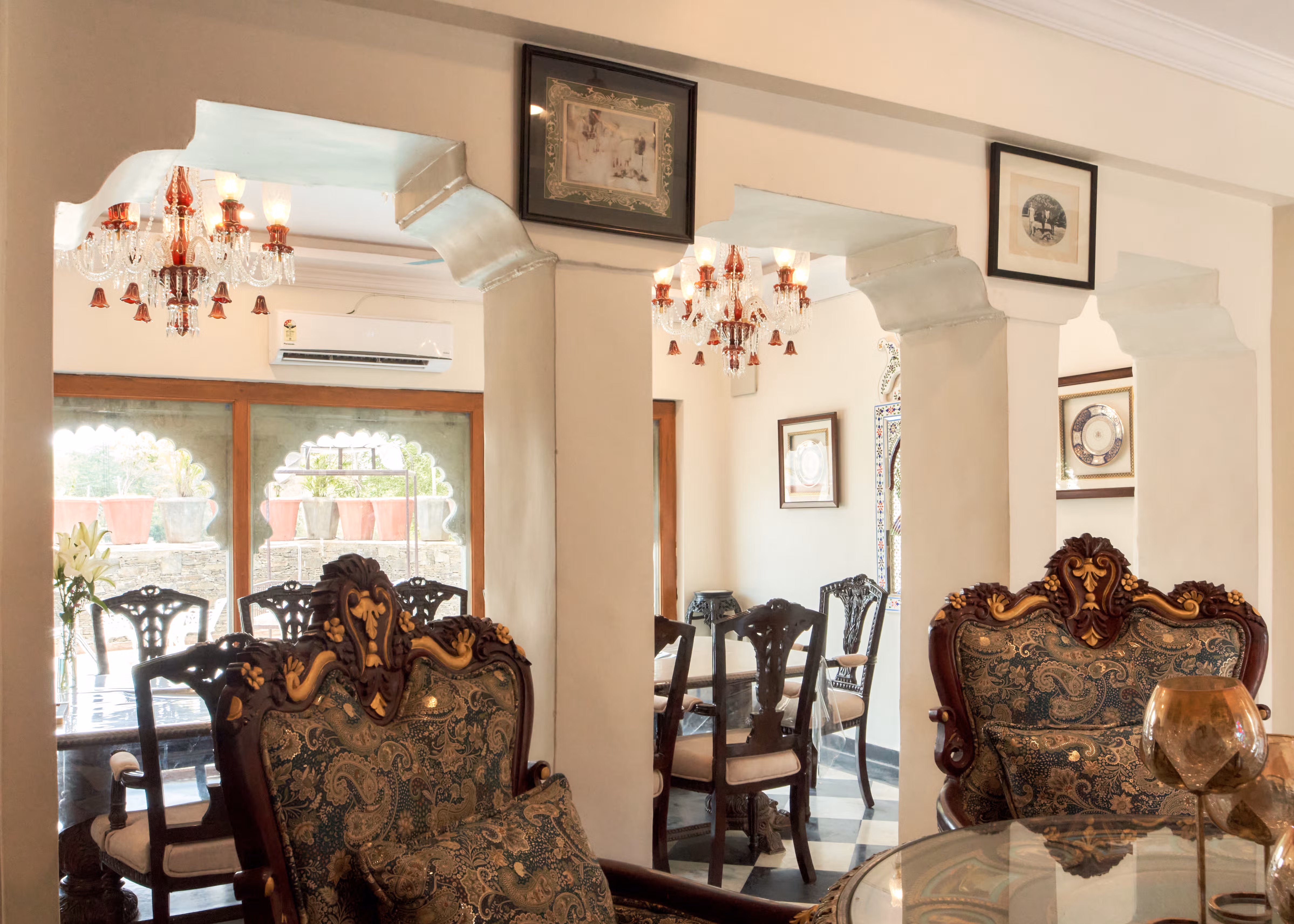 Carved upholstered chairs beside a columned opening into the dining room, with chandeliers and garden-facing windows.