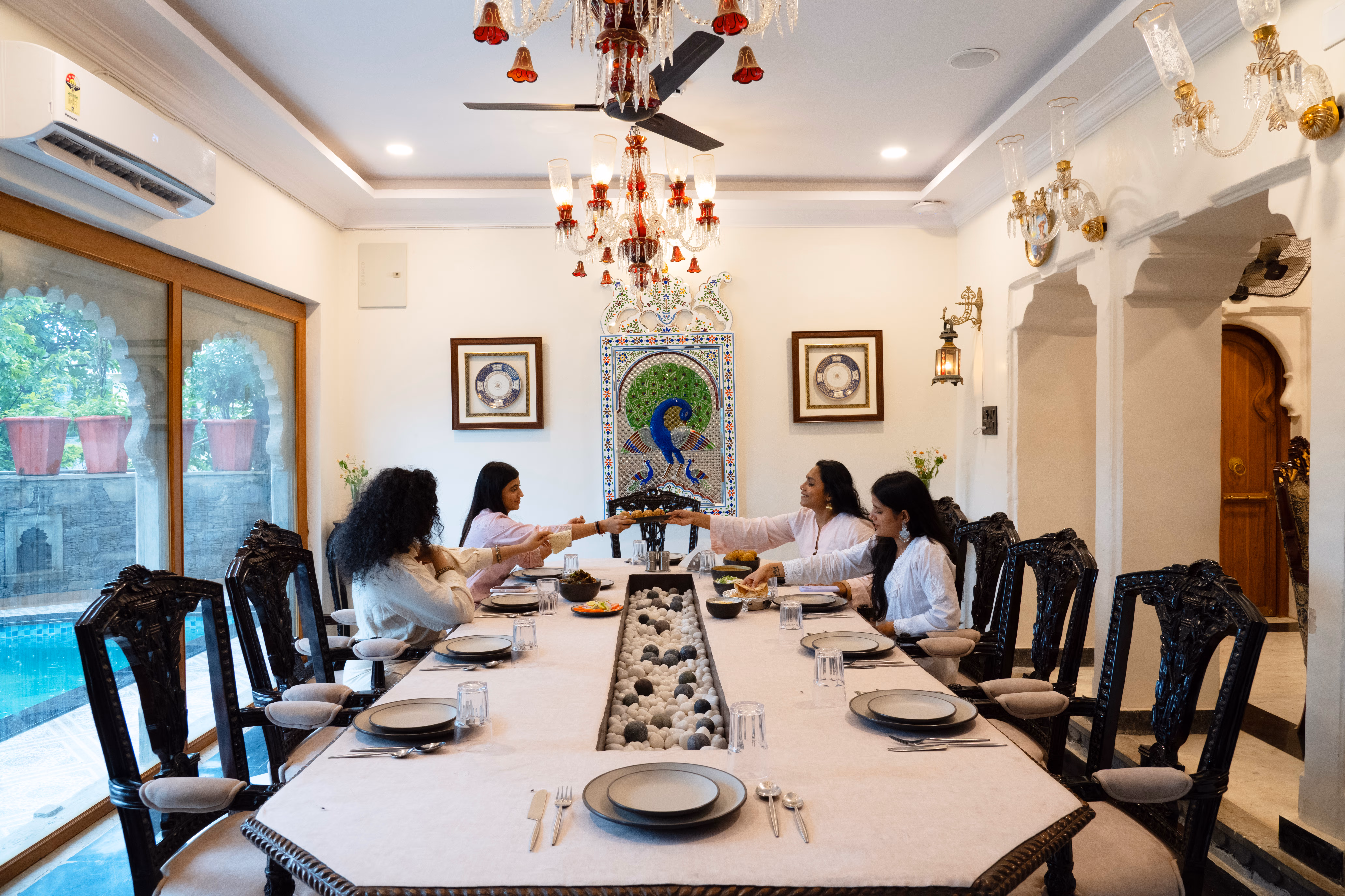 Guests dining at a long table beneath a chandelier, with carved chairs and large garden-facing windows.