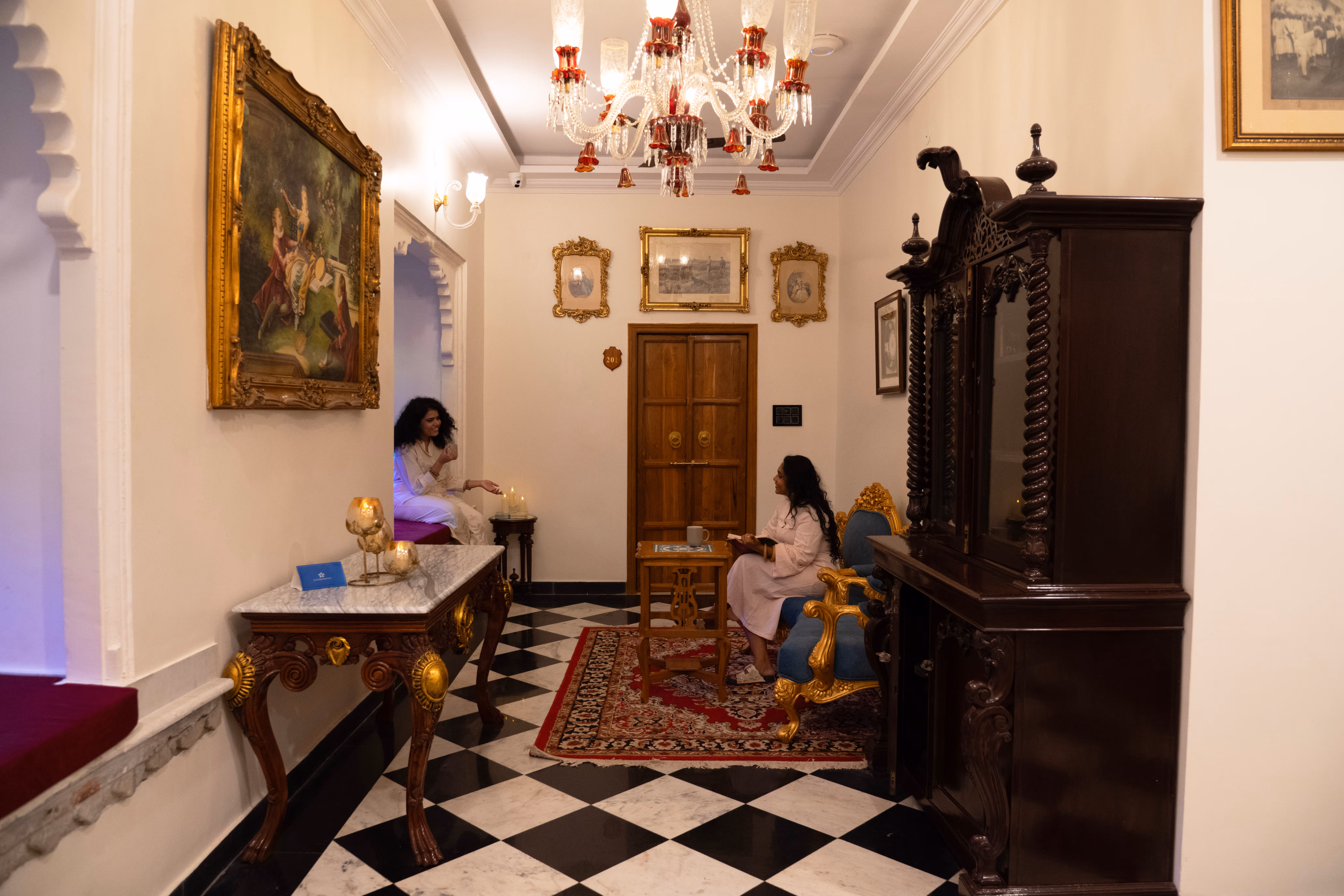Guests seated in a palace lounge with carved furniture, framed paintings and a black-and-white tiled floor.