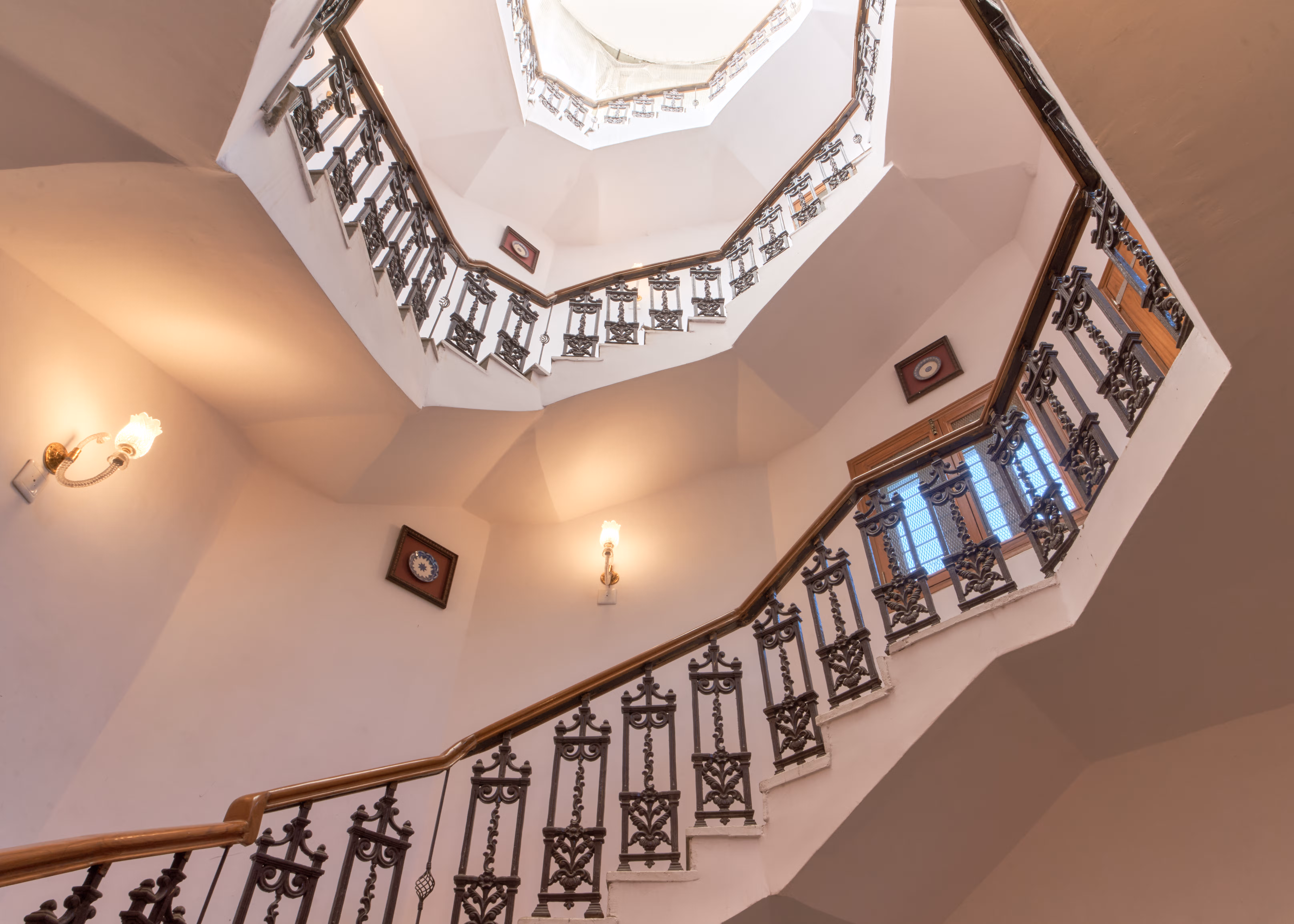 Upward view of a staircase with patterned metal balustrades, cream walls and framed pictures.